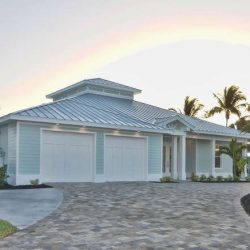 Single-story house with a metal roof, two garage doors, paved driveway, and palm trees in the landscaping.