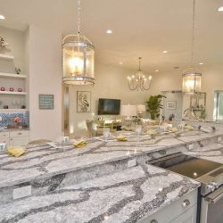 Modern kitchen with gray granite countertops, glass pendant lights, and a view into a bright, open living area.