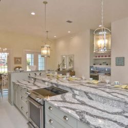 Modern kitchen with gray marble countertops, stainless steel appliances, hanging lights, and a dining area in the background.