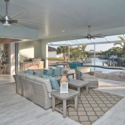 Covered patio with wicker furniture, ceiling fans, and views of a pool and waterway lined with palm trees.