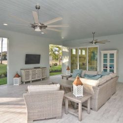 Covered patio with wicker furniture, ceiling fans, TV, and large windows overlooking a landscaped yard.