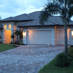 Single-story house with a tile roof, palm trees, brick driveway, and exterior lights on at dusk.