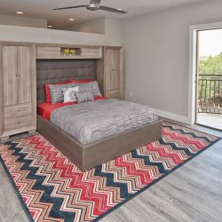 Modern bedroom with a bed on a zigzag patterned rug, built-in wooden storage, and an open door to a balcony.