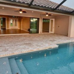 Covered patio with ceiling fans and sliding doors opening to a tiled area next to a rectangular pool in a residential home.