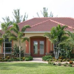 Single-story house with a red tile roof, tan walls, double front doors, and landscaped yard with palm trees.