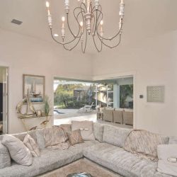 Modern living room with a light gray sectional sofa, chandelier, nautical decor, and large windows facing a patio and pool.