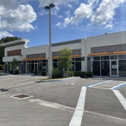 Empty storefronts with orange awnings in a small shopping center, viewed from a vacant parking lot on a sunny day.