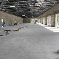 Large, unfinished commercial building interior with concrete floors, exposed steel ceiling, and workers in safety vests.