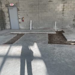 A concrete floor with two rectangular sections cut out, exposing soil and white pipes; gray cinderblock wall in background.