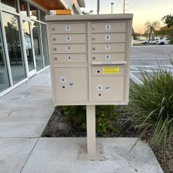 A beige outdoor cluster mailbox unit with numbered compartments stands on a concrete sidewalk near a building and parking lot.