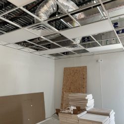 Suspended ceiling installation in progress with tiles, tools, and materials stacked on the floor of a partially finished room.