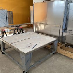 A large stainless steel table on wheels stands in front of an industrial metal machine in a workshop setting.