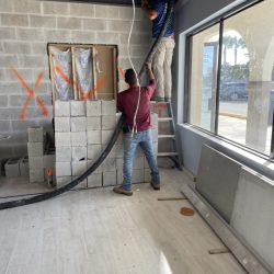 Two workers install wiring inside a partially constructed room with cinder block walls and exposed ceiling beams.