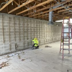 Construction worker in a yellow vest kneels near a cinder block wall under exposed ceiling beams and wiring.