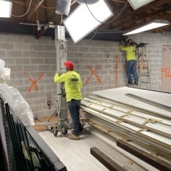 Two workers in safety gear install equipment and mark measurements on a concrete wall at a construction site.